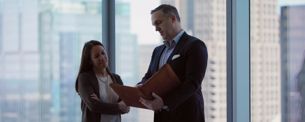 A feminine subject and a masculine subject in business attire, reviewing documents in a folio while in an office that is overlooking skyscrapers.