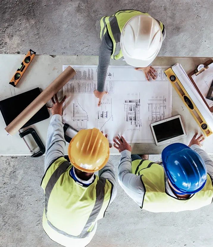 An overhead view of three people in construction hats and vests. They are looking at plans on a desk.