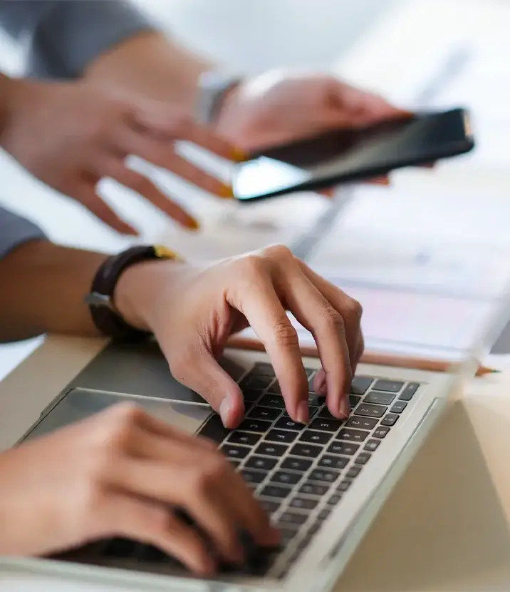 A photo of two people in business attire reviewing documents on a table. One has a phone, the other a laptop.